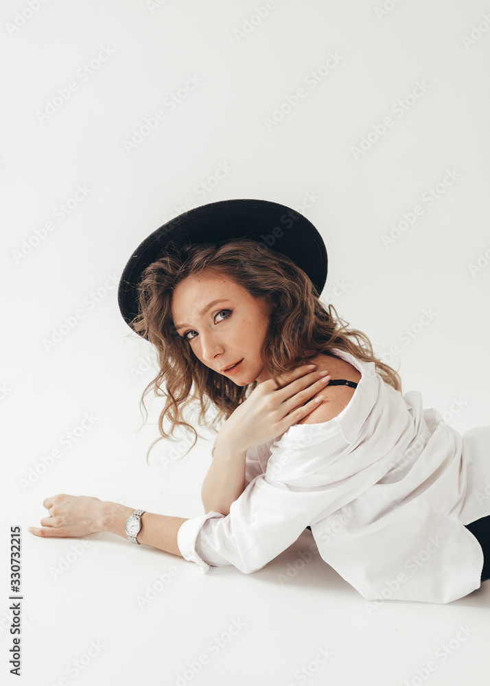 The concept of a trendy modern fashion trend shooting in a photo Studio , a beautiful sexy attractive gentle daring girl in a black hat, in black underwear, in a large shirt posing on a white backgrou The concept of a trendy modern fashion trend shooting in a photo Studio , a beautiful sexy attractive gentle daring girl in a black hat, in black underwear, in a large shirt posing on a white backgrou