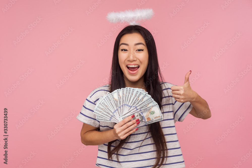 I like my rich life! Portrait of joyful angelic girl with nimbus over head showing thumbs up and holding money, satisfied with big income, lottery winning. studio shot isolated on pink background I like my rich life! Portrait of joyful angelic girl with nimbus over head showing thumbs up and holding money, satisfied with big income, lottery winning. studio shot isolated on pink background