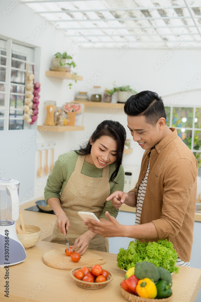 Beautiful young couple is using a cellphone and smiling while cooking in kitchen at home