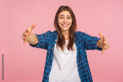Fototapeta Let me embrace you! Portrait of kind-hearted girl in checkered shirt opening her arms wide to hug you, welcoming with toothy smile, sharing love