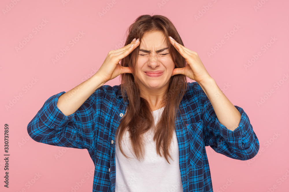Severe headache, intense migraine. Portrait of unhealthy girl in checkered shirt clutching head and grimacing from unbearable pain, stress tension in head. studio shot isolated on pink background Severe headache, intense migraine. Portrait of unhealthy girl in checkered shirt clutching head and grimacing from unbearable pain, stress tension in head. studio shot isolated on pink background