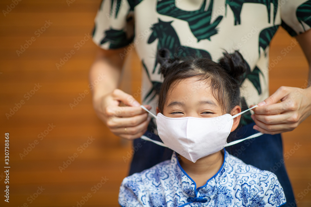 The parent is helping the Asian girl to wear the surgical mask for protection of virus or air pollution