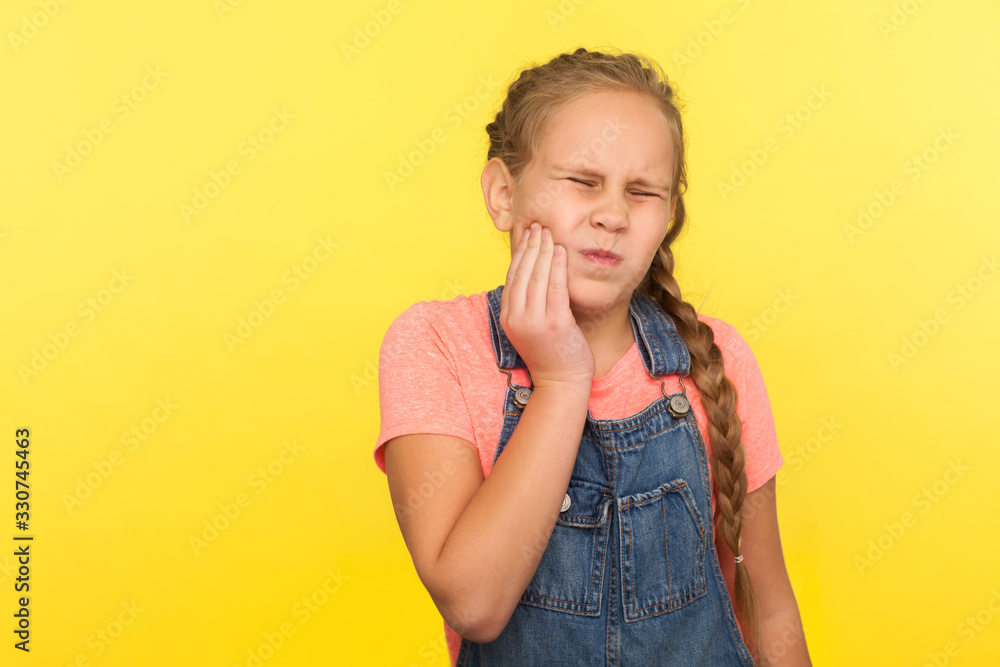 Dental pain in children. Portrait of unhealthy little girl with braid in denim overalls touching cheek, feeling severe toothache, oral health problems. indoor studio shot isolated on yellow background Dental pain in children. Portrait of unhealthy little girl with braid in denim overalls touching cheek, feeling severe toothache, oral health problems. indoor studio shot isolated on yellow background