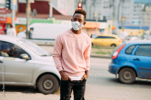 A young African man stands with a medical protective mask on his face at city street. © Stanislav