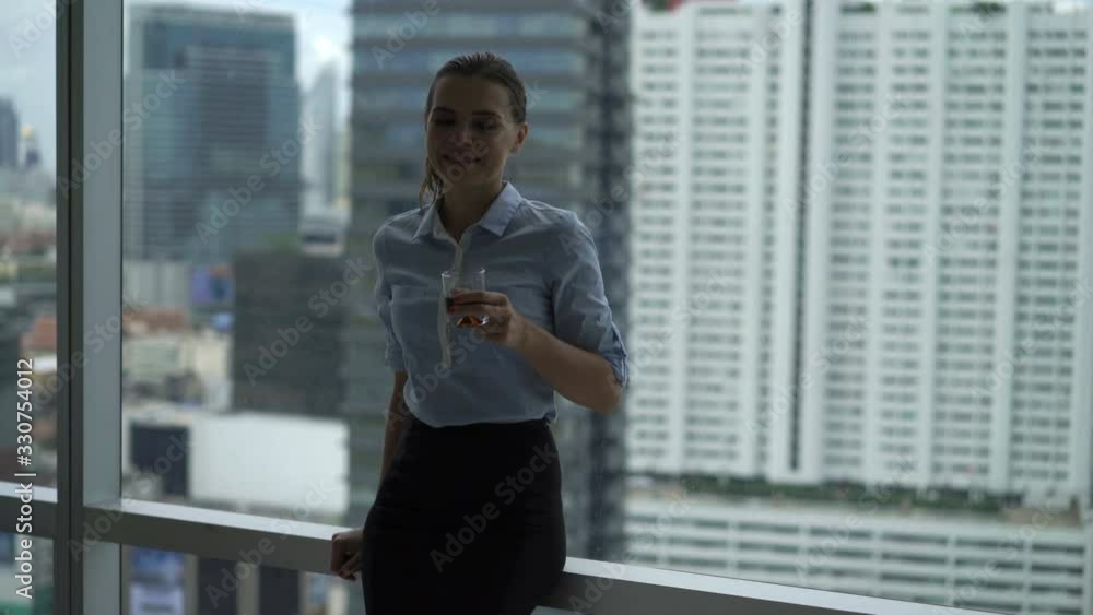 Young happy businesswoman drinking whiskey and relaxing by window at office Young happy businesswoman drinking whiskey and relaxing by window at office