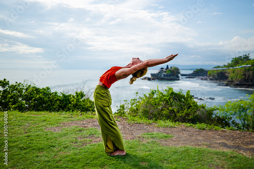 Young woman practicing yoga, standing in Hasta Uttanasana pose, deflection back. Outdoor yoga on the cliff. Yoga retreat. Tahan Lot temple Bali, Indonesia photo