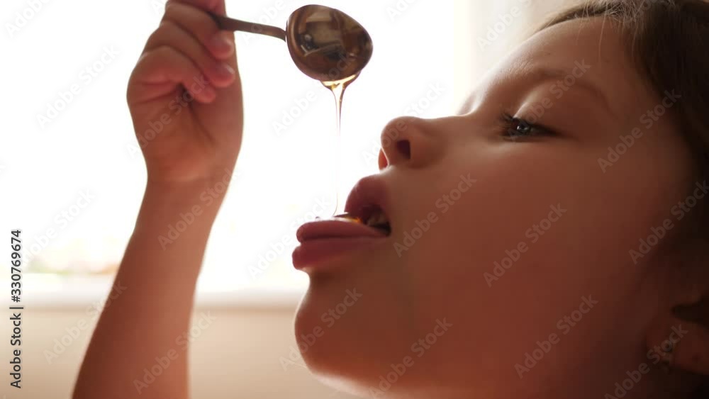 Little child girl eat honey as it drops on her tongue licking it out from the spoon