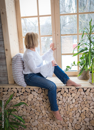 woman in white shirt and jeans. caucasian sitting window sill wooden and looking out the window, home interior