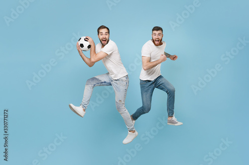 Excited two young men guys friends in white t-shirt isolated on pastel blue background in studio. Sport leisure lifestyle concept. Cheer up support favorite team with soccer ball jumping like running.