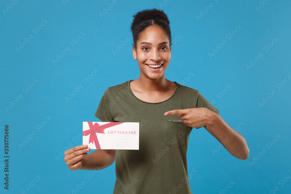 Excited young african american woman girl in casual t-shirt posing isolated on blue background studio portrait. People lifestyle concept. Mock up copy space. Pointing index finger on gift certificate. Excited young african american woman girl in casual t-shirt posing isolated on blue background studio portrait. People lifestyle concept. Mock up copy space. Pointing index finger on gift certificate.