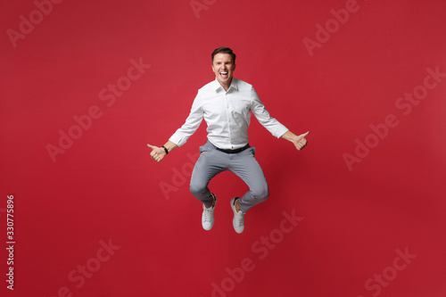 Crazy young business man in white shirt, gray pants posing isolated on bright red background studio portrait. Achievement career wealth business concept. Mock up copy space. Jumping showing thumbs up.