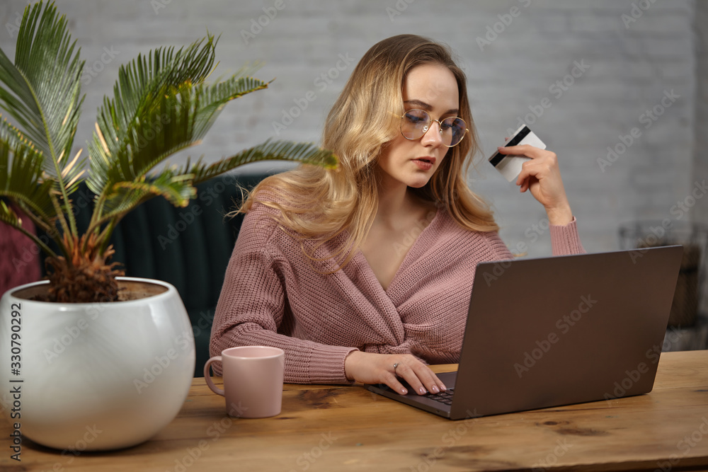 © nazarovsergey - Female in pink cardigan, glasses. Working on laptop and showing white plastic card. Sitting at wooden table with pink cup, palm in pot on it. Close up