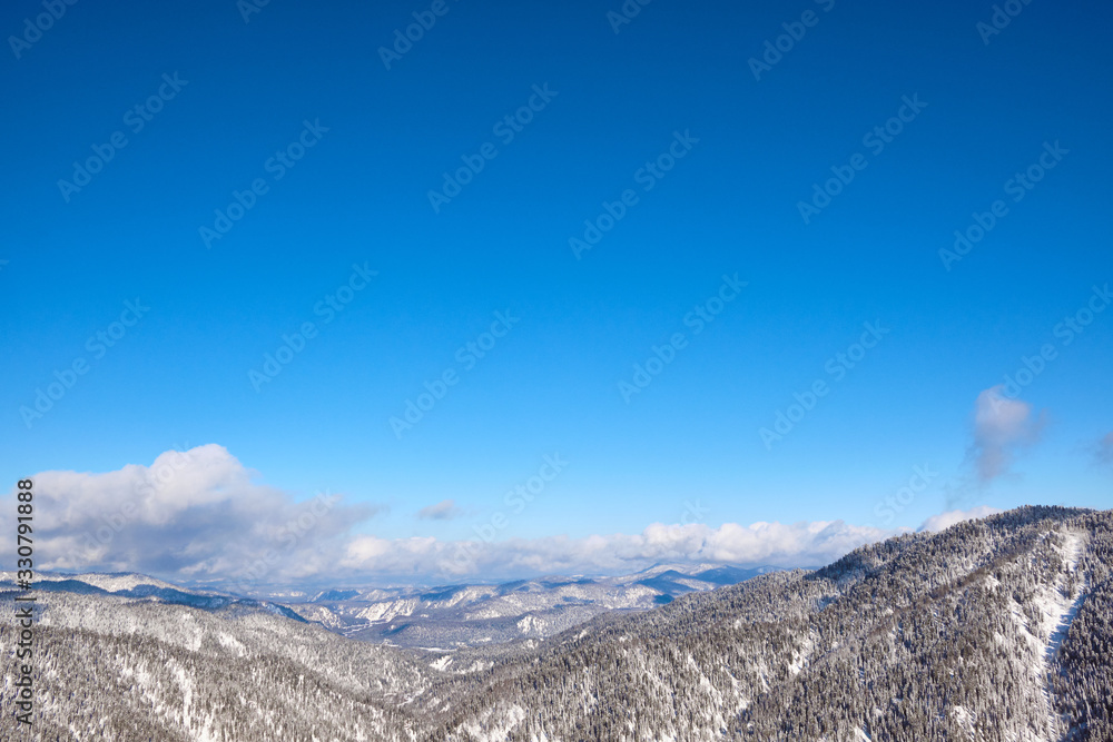 Winter mountain landscape. Snow-capped mountains overgrown with coniferous taiga against the background of clouds and blue sky. Russia. Altai Republic.