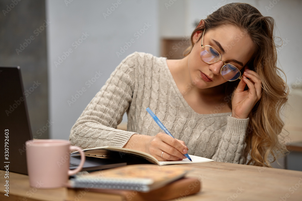 Blonde female blogger in glasses, beige sweater. Sitting in kitchen at wooden table, going to write something in her notebook. Close up Blonde female blogger in glasses, beige sweater. Sitting in kitchen at wooden table, going to write something in her notebook. Close up