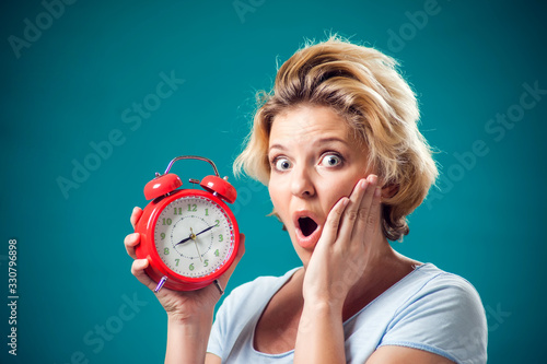 A portrait of woman holding alarm clock and feeling shocked because of oversleep