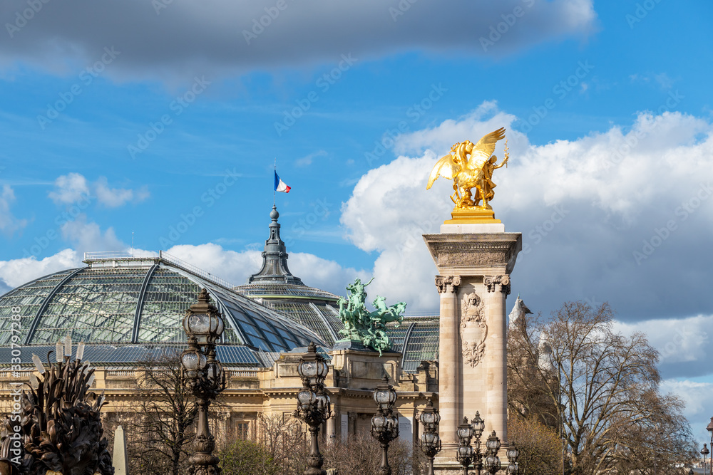 Golden statue and lanterns of Pont Alexandre III bridge with Grand Palais and French flag waving on top of the building - Paris, France