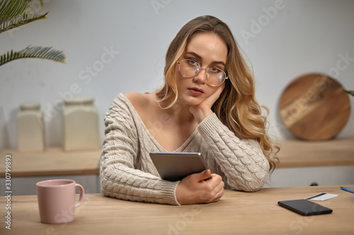 Lady blogger in glasses, beige sweater. Holding tablet, sitting in kitchen at wooden table with smartphone, plastic card and pink cup on it. Close-up