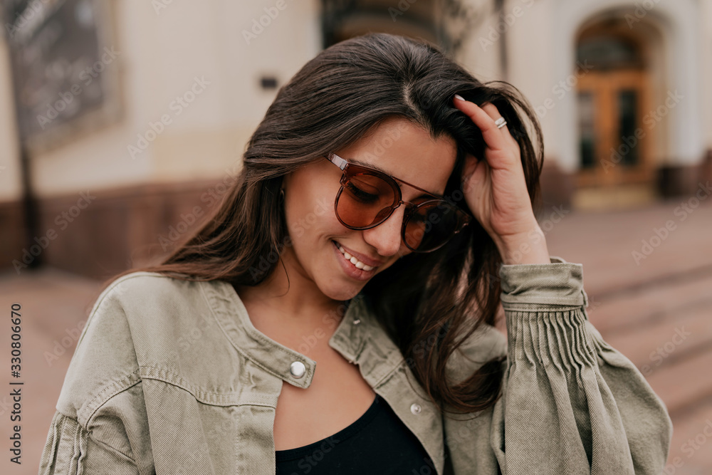 Charming shy european woman with dark hair wearing sunglasses wearing jacket walking on the city in good sunny day Charming shy european woman with dark hair wearing sunglasses wearing jacket walking on the city in good sunny day