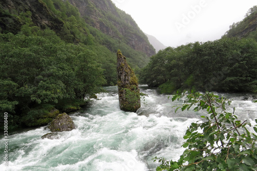 A tall rock standing in the rapid river of the Flamselvi surrounded by woods and mountains between Myrdal and Flams next to the Rallarsvegen and the Flamsbana; Norway, Europe photo