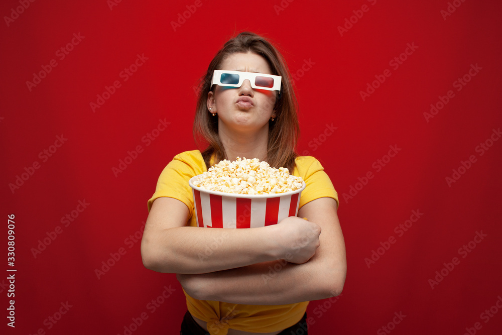 surprised young girl student watching a movie in 3D glasses and eating popcorn on a red background