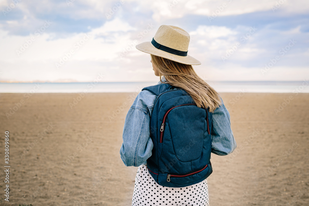 Young girl with backpack and hat looks at the horizon from the beach.