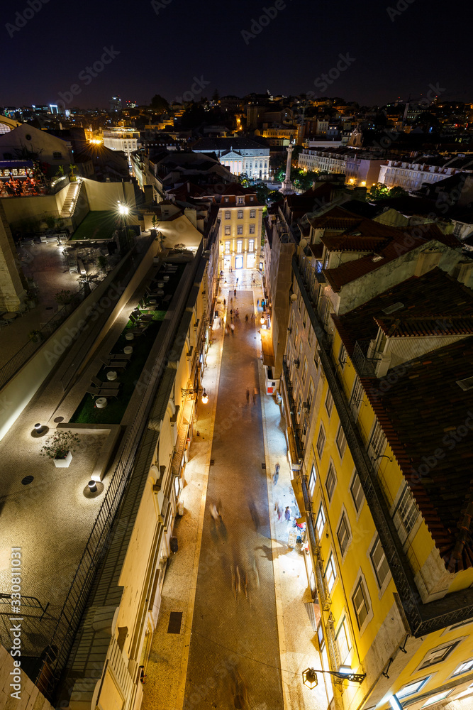 View of lit buildings and people on the Rua do Carmo street towards Rossio Square at the Baixa district in Lisbon, Portugal, from above at night.