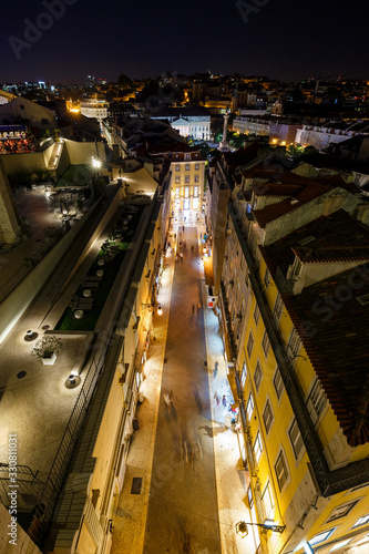 View of lit buildings and people on the Rua do Carmo street towards Rossio Square at the Baixa district in Lisbon, Portugal, from above at night.
