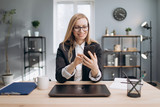 Cheerful lady with blond hair in eyeglasses using smartphone to solve business issues online. Mature woman in formal clothing smiling and sitting at modern office.