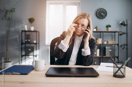 Happy mature lady with blond hair taking off eyeglasses after hard working day at office. Business woman in formal clothing talking on smartphone while sitting at table.