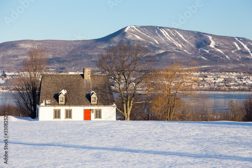 Beautiful early morning winter view of patrimonial white wooden house with steep shingled roof in rural setting with trees, river and mountains, St. Pierre, Island of Orleans, Quebec, Canada
