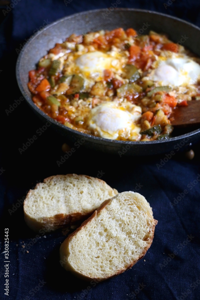 Shakshuka in a pan, with seasonal vegetables and eggs, and two slices of bread. Selective focus.