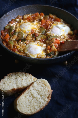 Shakshuka in a pan, with seasonal vegetables and eggs, and two slices of bread. Selective focus.