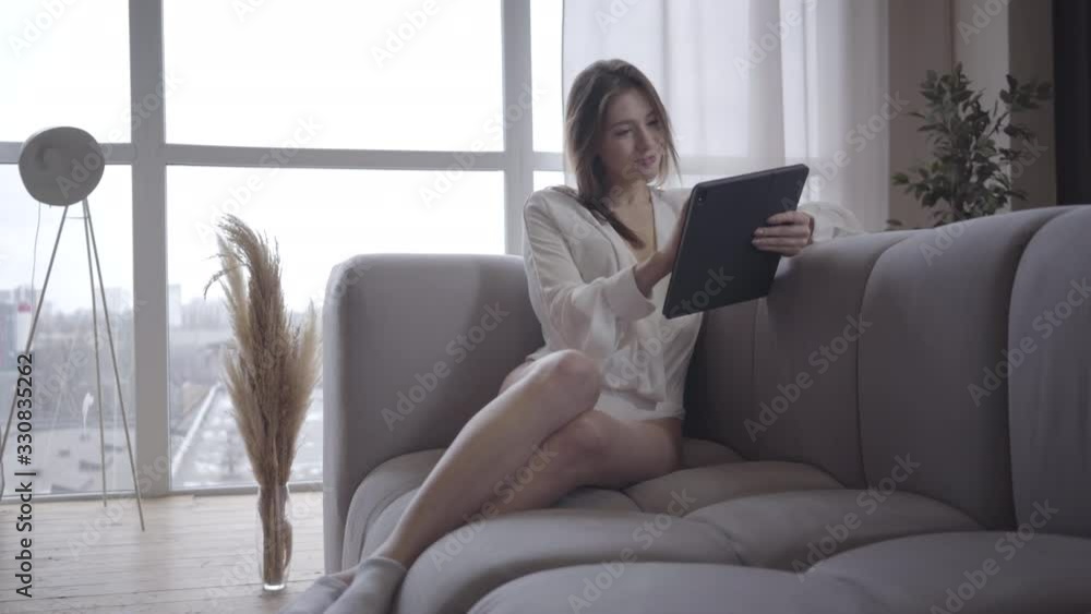 Charming smiling Caucasian girl sitting on couch and using tablet. Portrait of cheerful young woman using social media at home in the morning. Joy, lifestyle, wireless technology. Charming smiling Caucasian girl sitting on couch and using tablet. Portrait of cheerful young woman using social media at home in the morning. Joy, lifestyle, wireless technology.