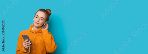 Ginger caucasian woman with freckles is listening cheerfully to musing using a phone and headphones while posing on a blue wall with free space