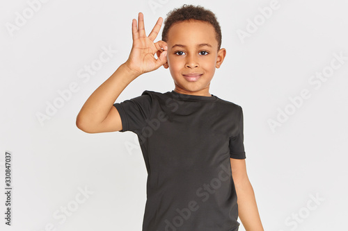 Body language. Portrait of friendly looking positive dark skinned little boy in t-shirt connecting fore finger and thumb making approval gesture, showing okay sign, saying Everything is fine photo