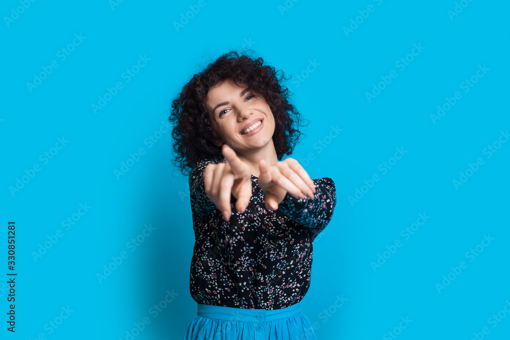 Joyous caucasian woman with curly hair is pointing to camera while posing on a blue background Joyous caucasian woman with curly hair is pointing to camera while posing on a blue background
