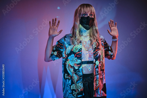 Stressful looking young adult woman posin in a neon studio in front of wuhan coronavirus projection, wearing shirt, medical mask and sportive bra holding her hands up