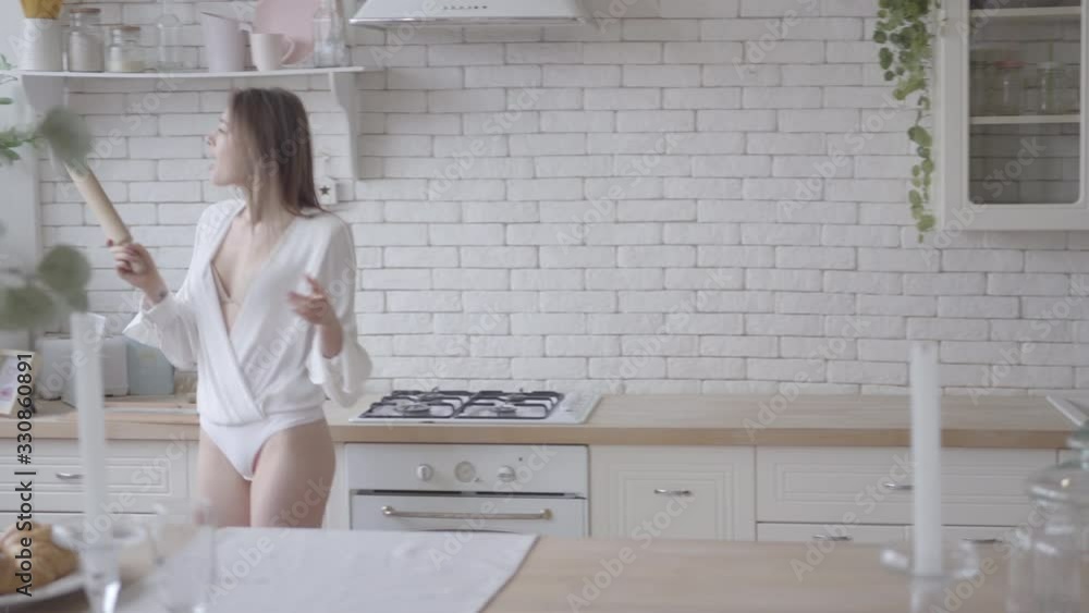 Joyful young Caucasian woman coming into modern kitchen, taking rolling pin and singing into it. Positive brunette girl having fun at home. Lifestyle, joy, happiness, leisure. Joyful young Caucasian woman coming into modern kitchen, taking rolling pin and singing into it. Positive brunette girl having fun at home. Lifestyle, joy, happiness, leisure.
