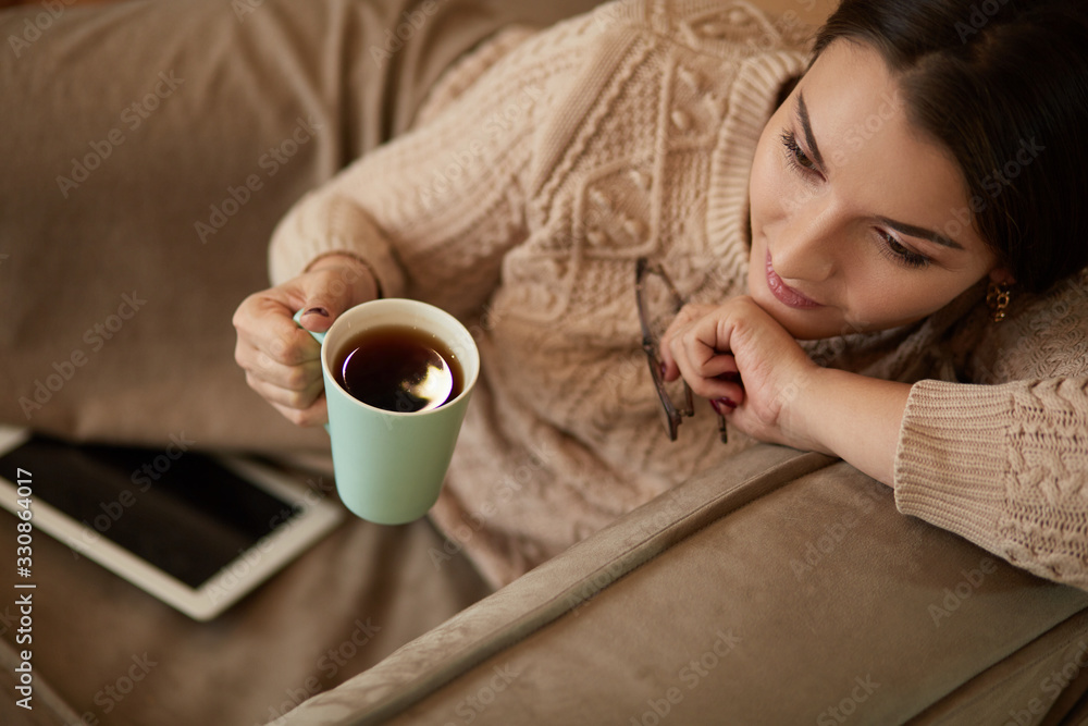Woman in home clothes sitting on the carpet near the sofa using a tablet , drinking coffee from a green cup Woman in home clothes sitting on the carpet near the sofa using a tablet , drinking coffee from a green cup