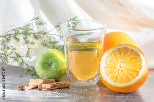 Vitamin lemon yellow tangerine peel drink in a transparent glass. Cinnamon and sliced orange on a wooden table. Juniper sprig apple