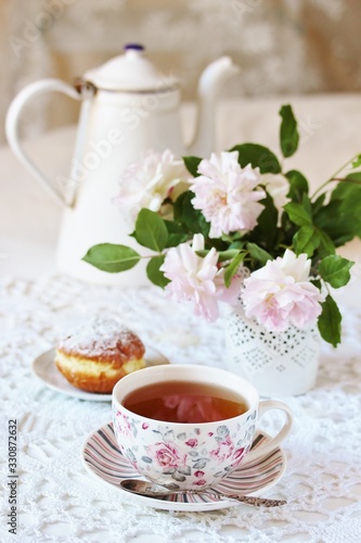 A tea party in the style of Shabby Chic. tea in the beautiful tea steam with a pattern "rose". Delicate pink roses in a vase, white kettle in retro style. On a white lace tablecloth. Soft focus.