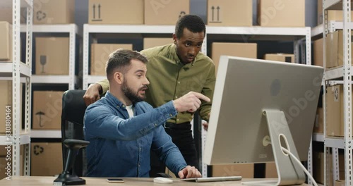 Caucasian young postman sitting ar table with computer and African American stannding beside while they talking and working. Cheerful men of delivery work discussing something and watching on screen. photo