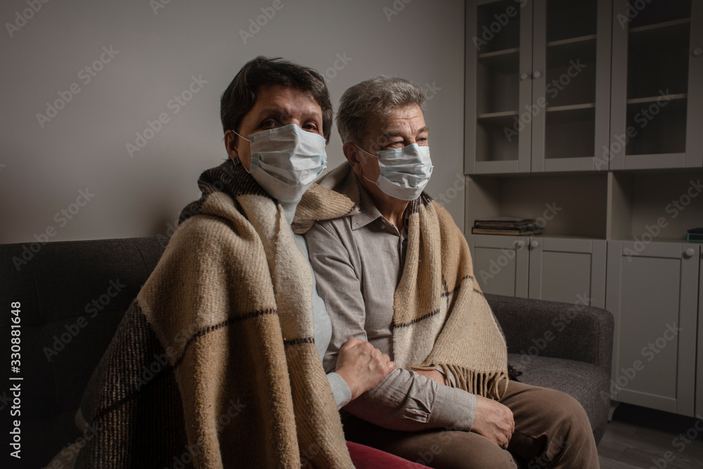 A couple sitting on a sofa wearing a protective mask. An infectious agent protection gear including a mask. An sick older man and woman wearing protective masks to protect against virus. Coronavirus.