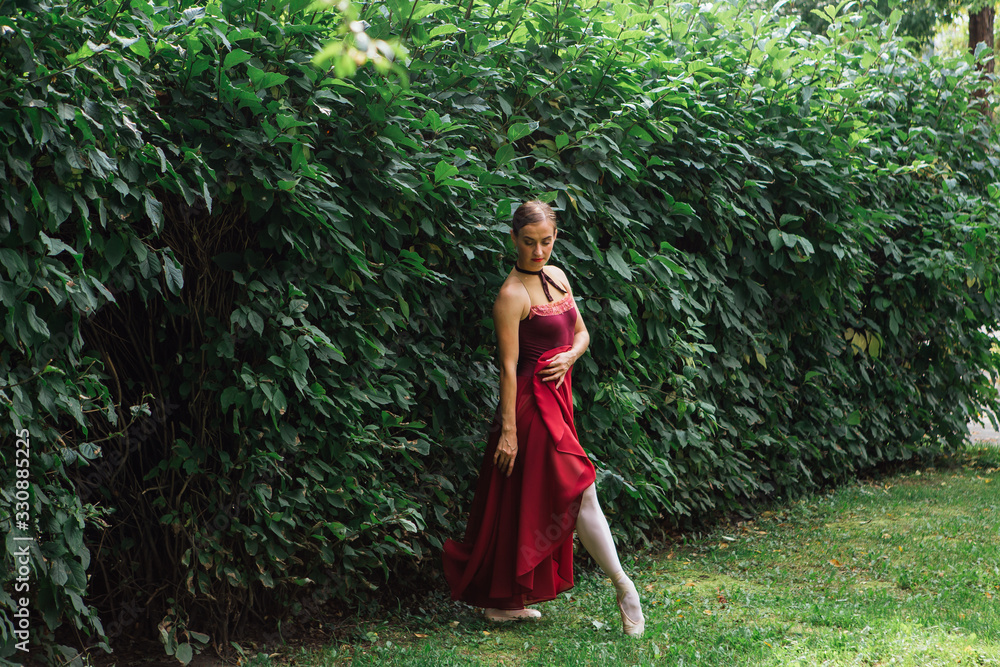 Woman ballerina in red ballet dress dancing in pointe shoes posing next to the wall of bushes Woman ballerina in red ballet dress dancing in pointe shoes posing next to the wall of bushes