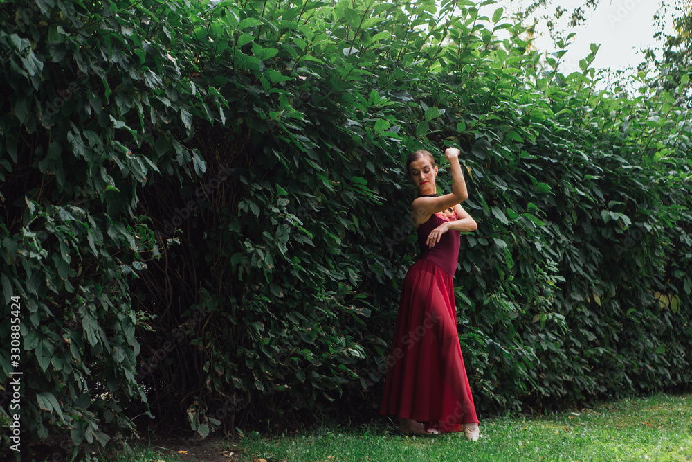 Woman ballerina in red ballet dress dancing in pointe shoes posing next to the wall of bushes Woman ballerina in red ballet dress dancing in pointe shoes posing next to the wall of bushes