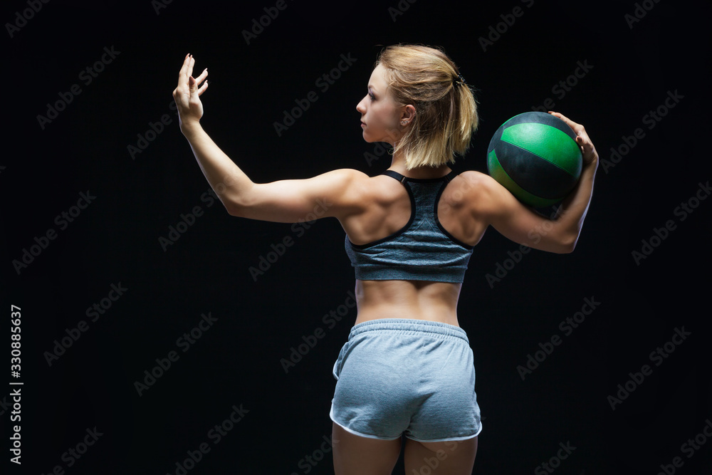 Back view of strong sporty girl in top and jean shorts showing muscles and holding a ball on black background Back view of strong sporty girl in top and jean shorts showing muscles and holding a ball on black background