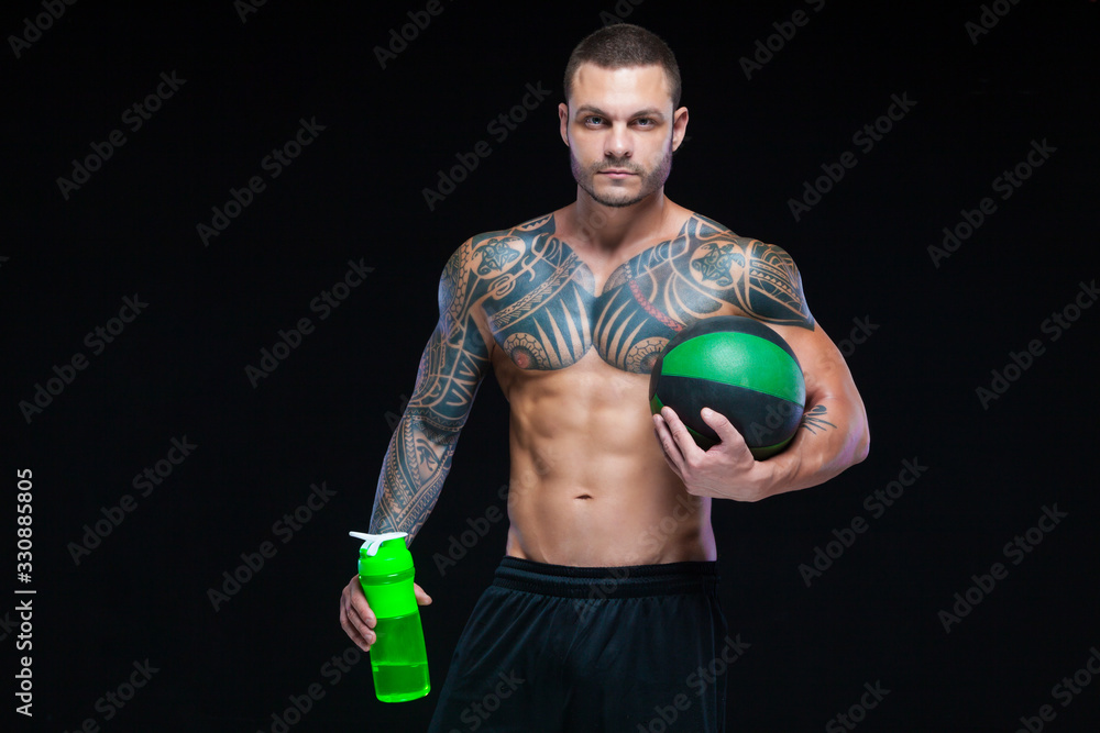 Muscular man bodybuilder with tattoos. Man posing on a black background with water bottle and ball