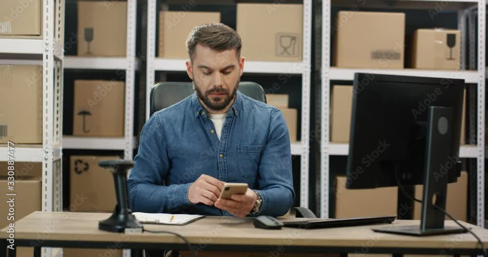 Caucasian man talking on mobile phone while working at post office store with parcels and on computer. Postman registering box and filling in invoice while speaking on cellphone in mail storage. Caucasian man talking on mobile phone while working at post office store with parcels and on computer. Postman registering box and filling in invoice while speaking on cellphone in mail storage.