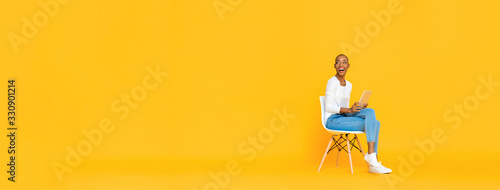 Trendy smiling African American woman sitting on a chair using tablet computer thinking and looking at empty space aside isolated yellow banner background