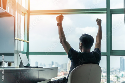 Business achievement concept with happy businessman relaxing in office or hotel room, resting and raising fists with ambition looking forward to city building urban scene through glass window photo